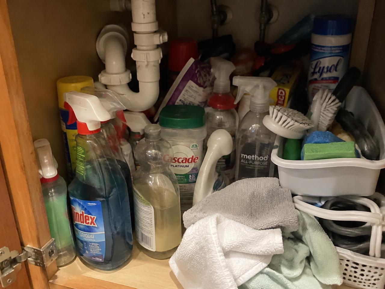 Cluttered under-sink cabinet with bottles stacked in front of each other around a drain pipe